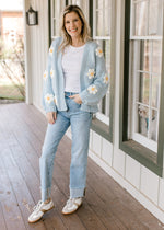 Model wearing sneakers, jeans and a knit pale blue cardigan with white daisies and long sleeves. 