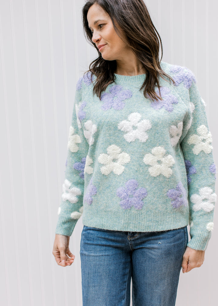Model wearing a cozy blue-green sweater with lavender and cream textured flowers and long sleeves.
