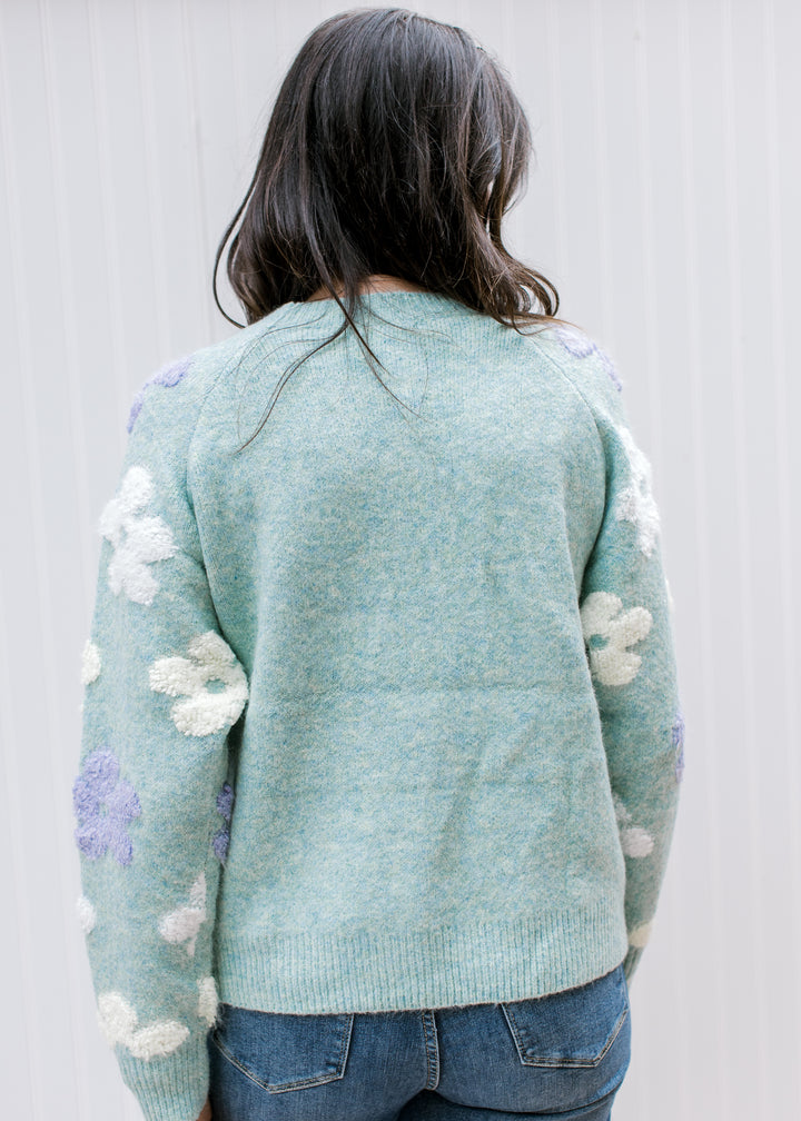 Back view of model wearing a blue green sweater with flowers, a round neckline and long sleeves. 