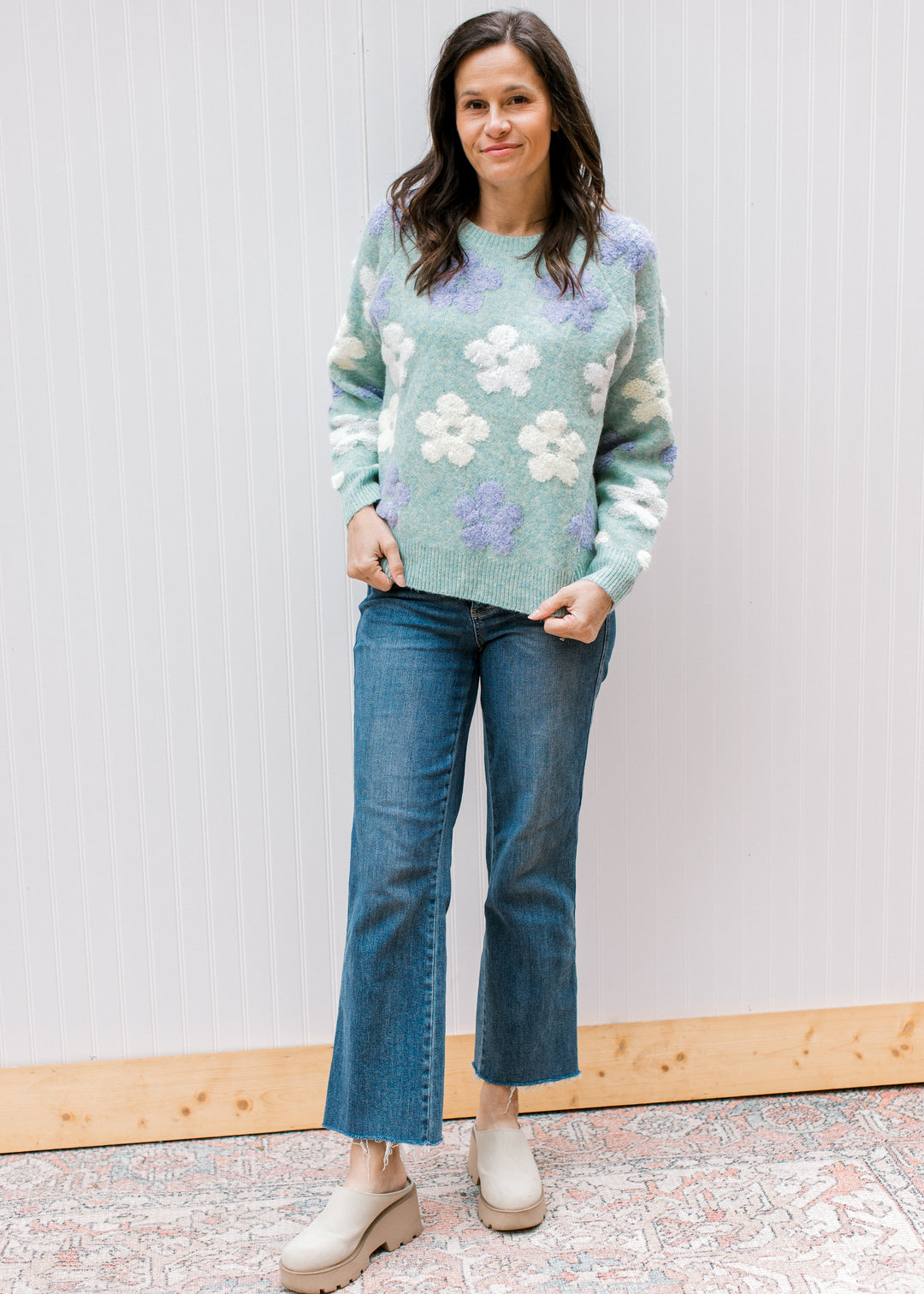 Model wearing jeans, mules and a blue green sweater with textured flowers and long sleeves. 