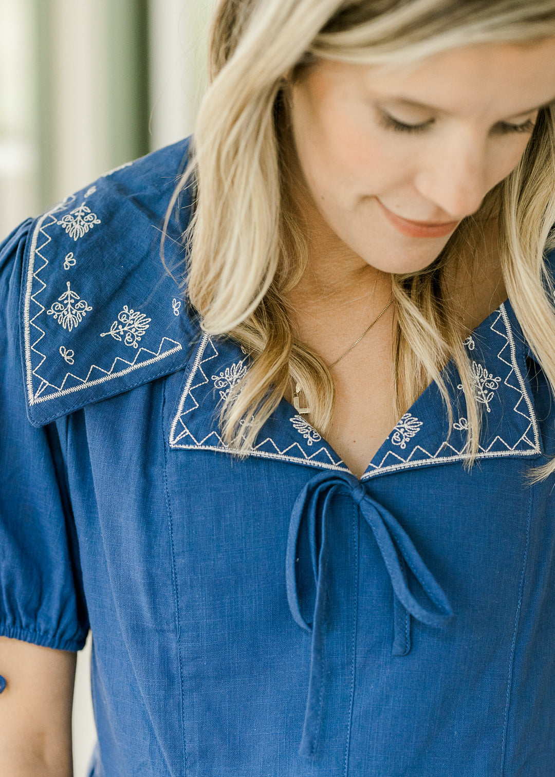 Model wearing a blue top with cream embroidery on exaggerated collar and hem. 