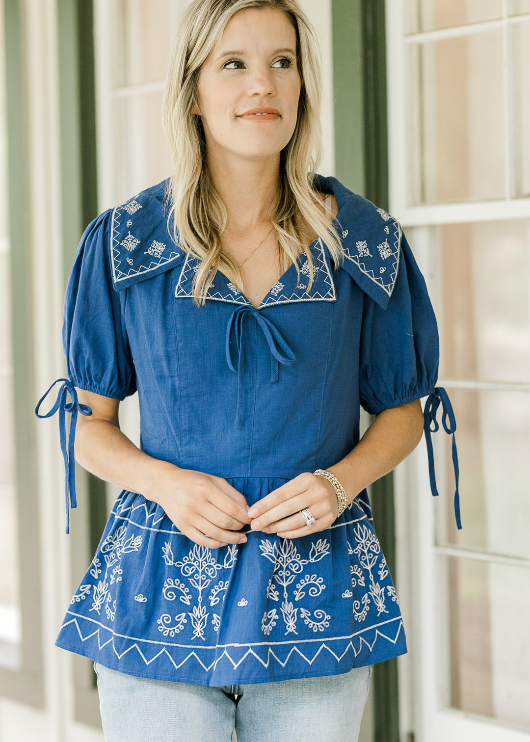 Model wearing a blue babydoll top with cream floral and tie at neck and sleeve. 