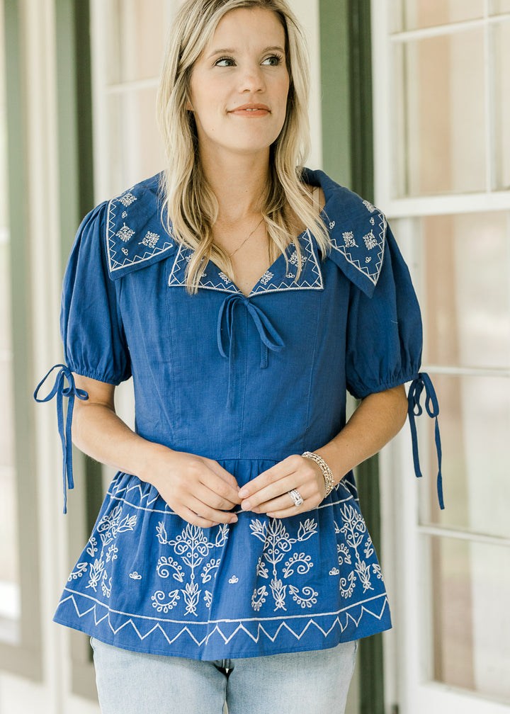 Model wearing a blue babydoll top with cream floral and tie at neck and sleeve. 