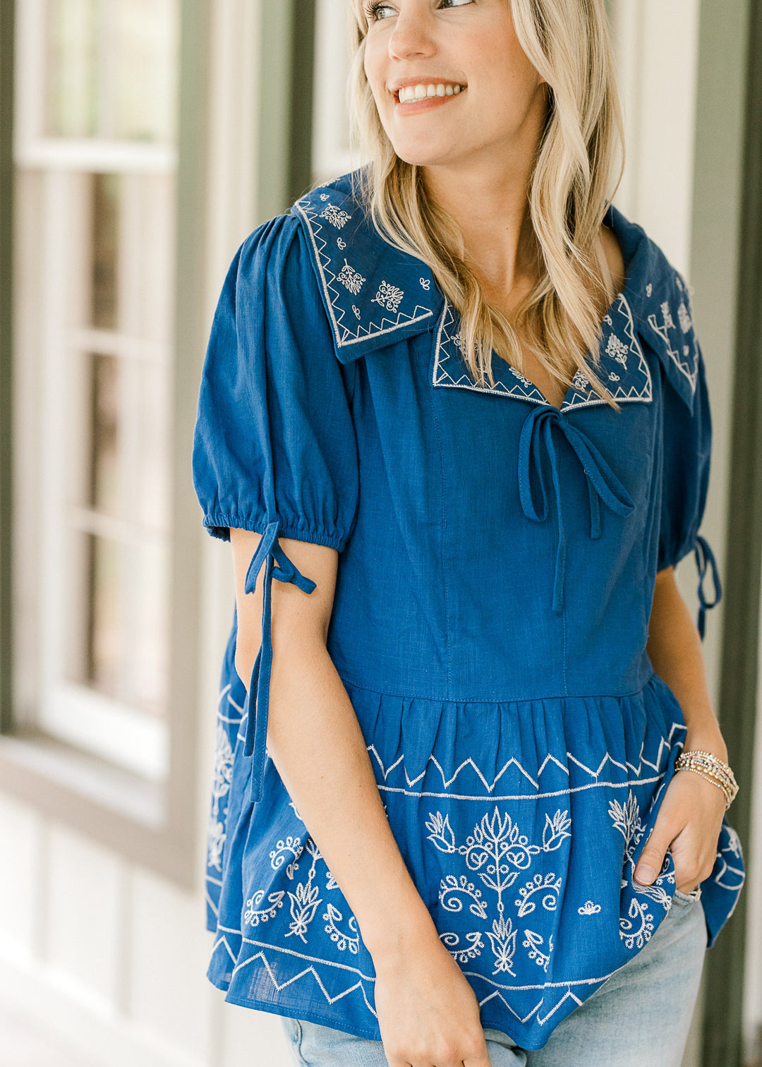 Woman wearing a blue top with cream embroidery with a blurred background. 