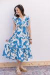 Model wearing sandals and a bright blue midi with cream floral, ruffle cap sleeves and side ties. 