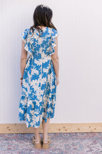 Back view of Model wearing a bright blue midi with cream floral, ruffle cap sleeves and a v-neck.