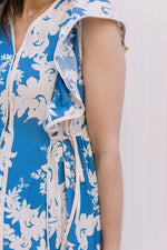 Close up of side tie at waist and ruffle cap sleeve on a bright blue midi with cream floral. 