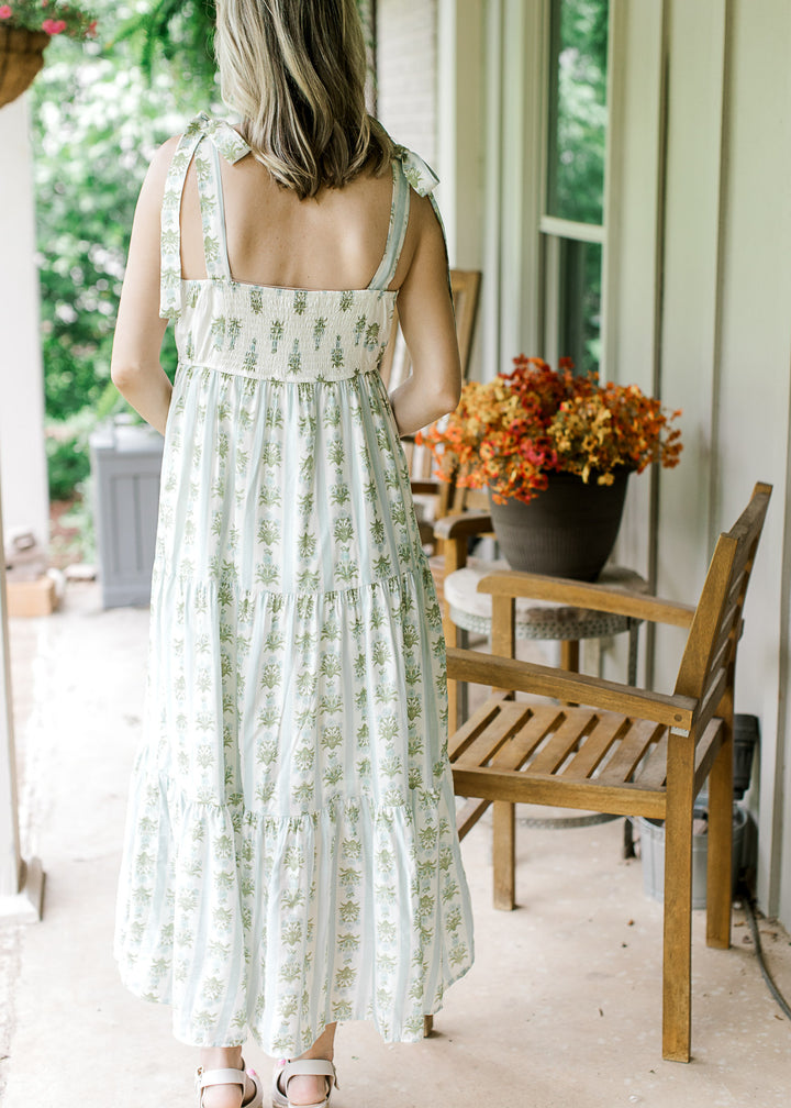 Back view of Model wearing a sleeveless cream midi with blue and green striped floral, smocked back.