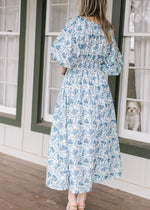 Back view of Model wearing a cream midi with blue floral, smocked waist and 3/4 bubble sleeves.