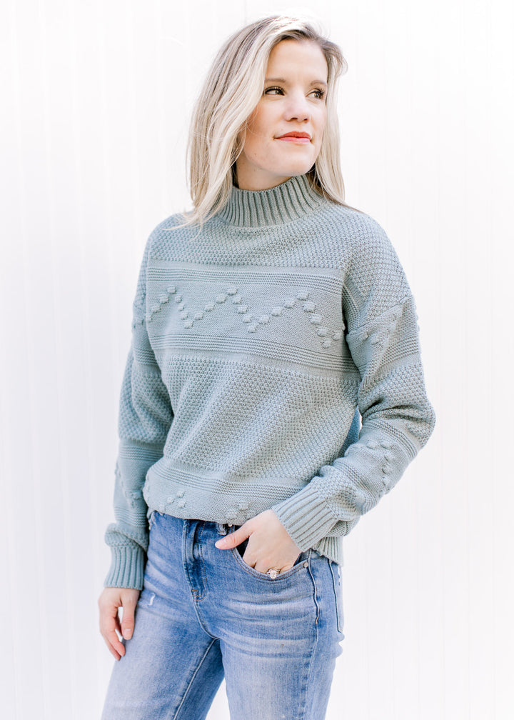 Model wearing a blue green sweater with a mock neckline, long sleeves and ribbed detailing. 