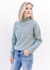Model wearing a blue green sweater with a mock neckline, long sleeves and ribbed detailing. 