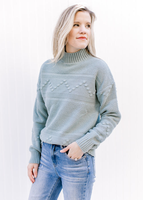 Model wearing a blue green sweater with a mock neckline, long sleeves and ribbed detailing. 