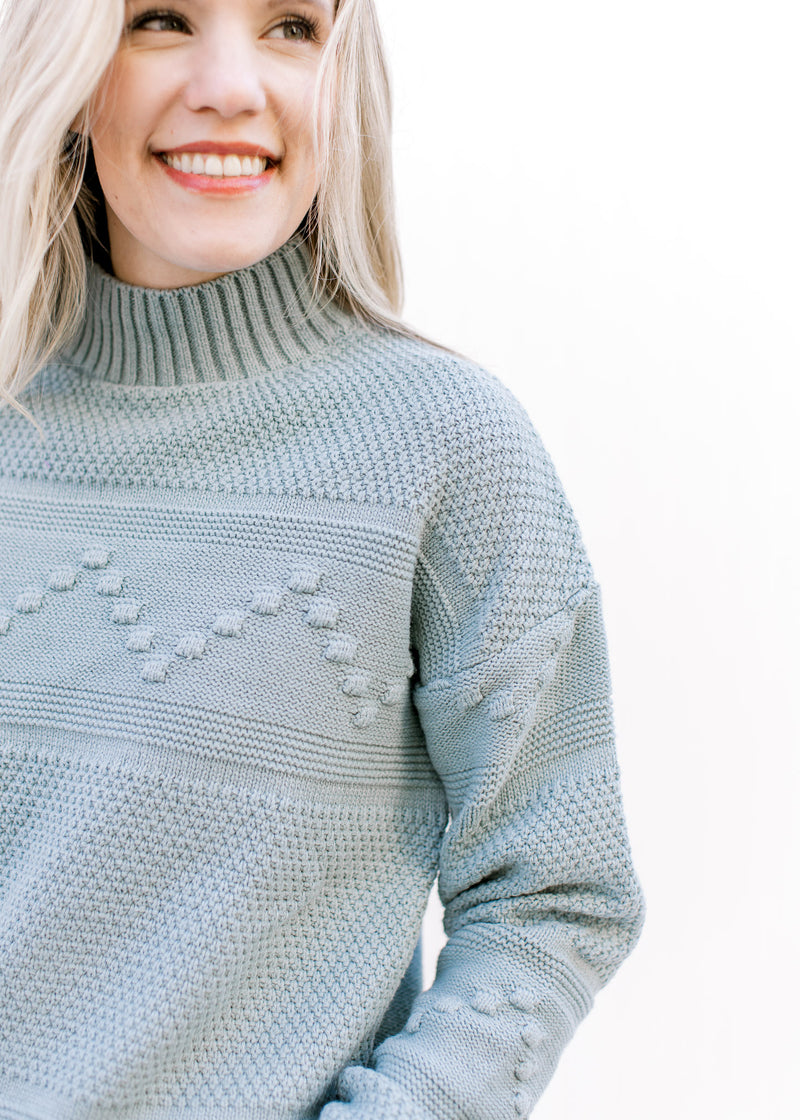 Close up of mock neck and Pom pom detail on a blue green sweater with long sleeves.