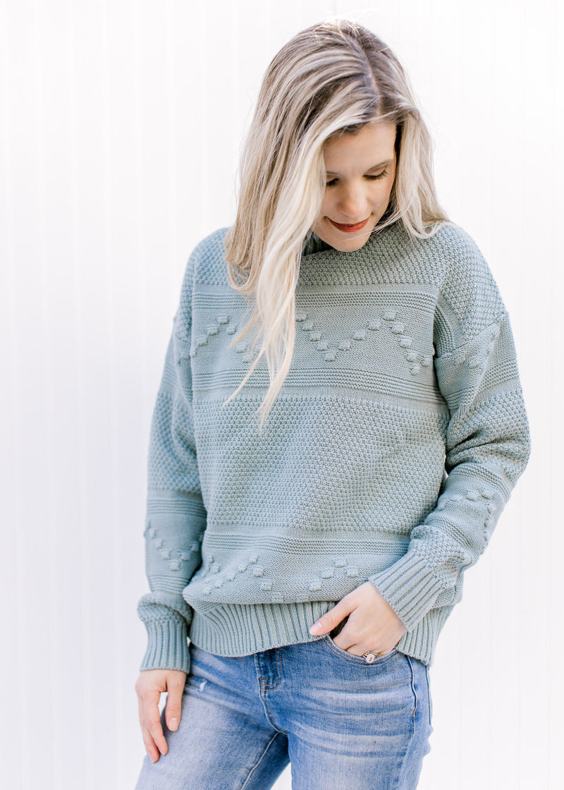 Model wearing jeans and a blue green sweater with a mock neck, long sleeves and ribbed detailing. 
