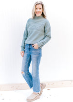 Model wearing jeans, mules and a blue green sweater with a mock neck and long sleeves. 