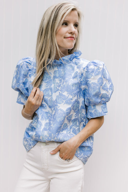 Model wearing a cream top with blue floral pattern, ruffle mock neck and short puff sleeves.