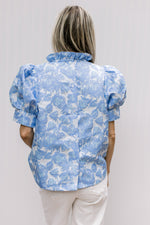 Back view of Model wearing a cream top with blue floral pattern, ruffle neck and short sleeves.