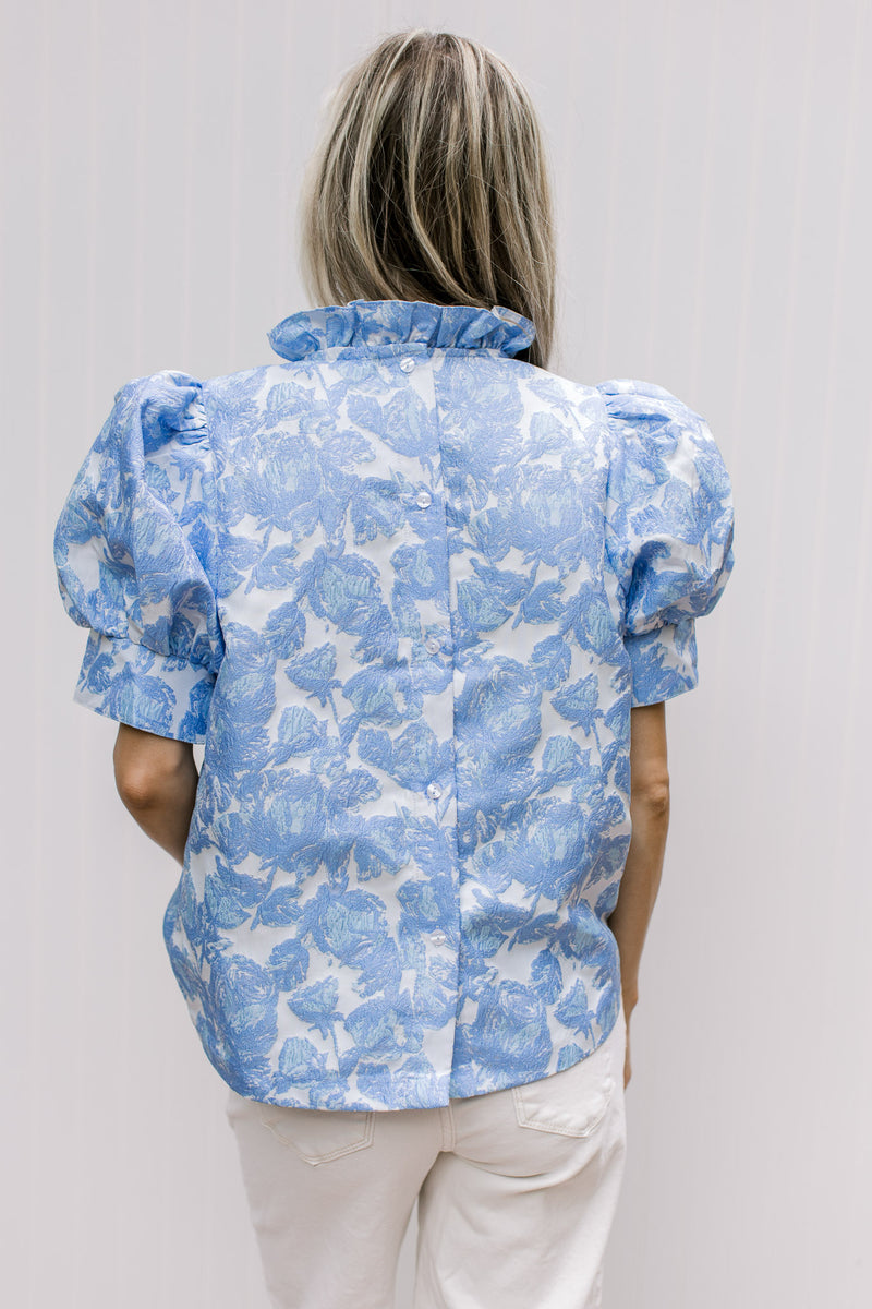 Back view of Model wearing a cream top with blue floral pattern, ruffle neck and short sleeves.