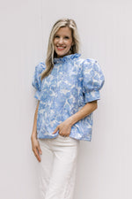 Model wearing white jeans with a cream top with blue floral, ruffle neck and short puff sleeves.