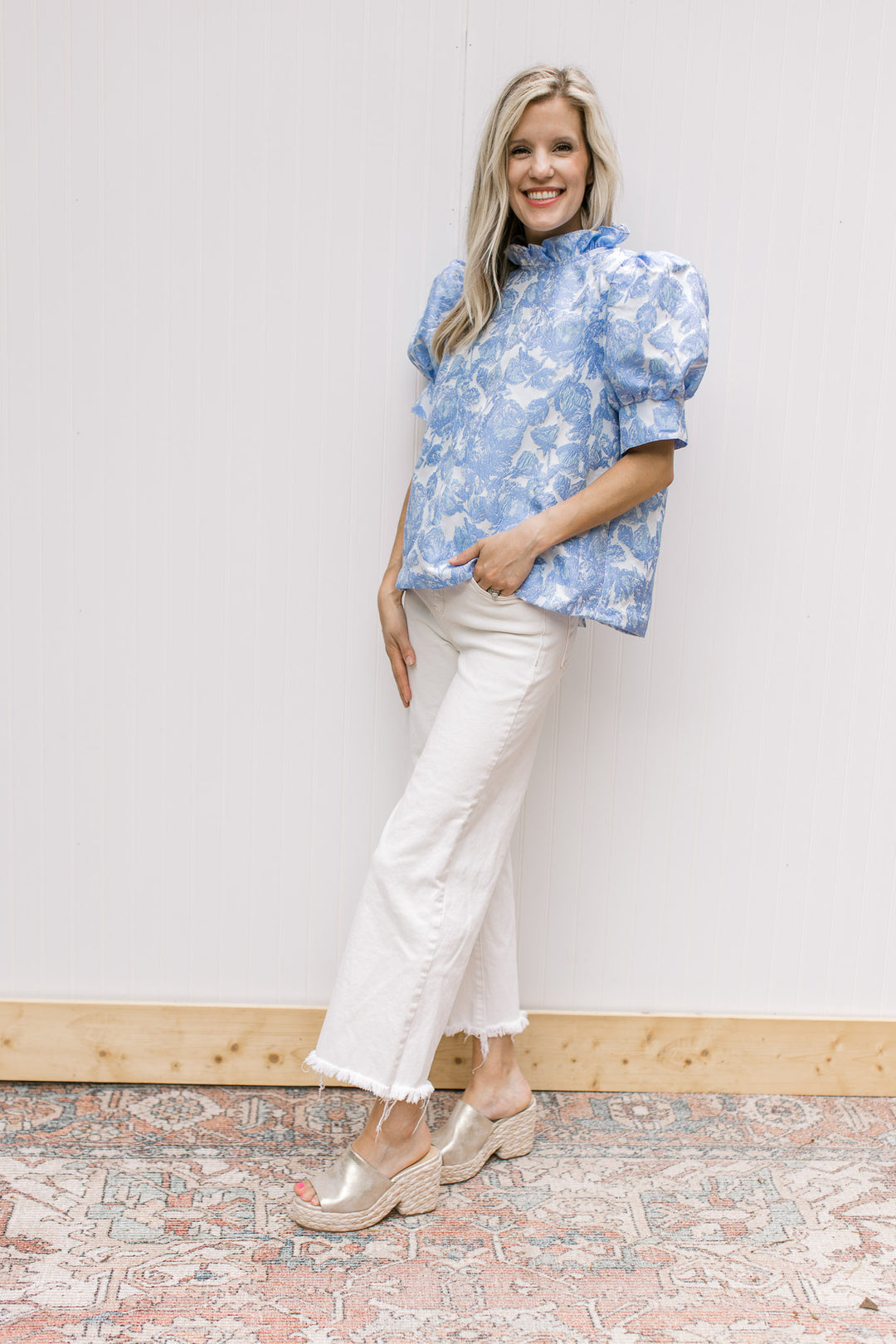 Model wearing white jeans, wedges and a cream top with blue floral pattern and ruffle neck.