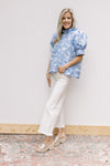 Model wearing white jeans, wedges and a cream top with blue floral pattern and ruffle neck.