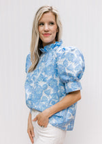 Model wearing a cream top with blue floral pattern, ruffle neck and short puff sleeves.