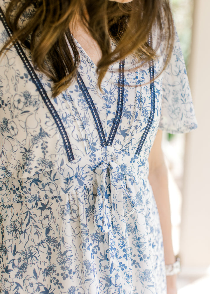 Close up of v-neck with double ladder trim and tie at waist of a cream midi with blue floral. 