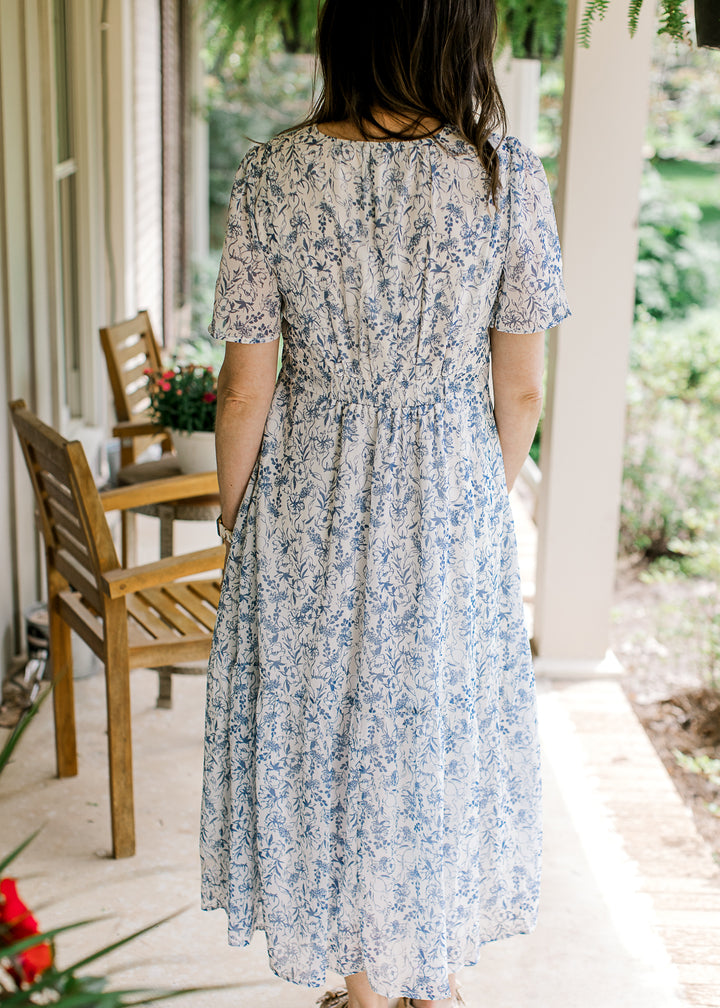 Back view of Model wearing a white midi with blue floral, flutter short sleeves and tie waist.