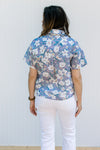 Back view of Model wearing a lavender top with flowers, short sleeves and a collar.