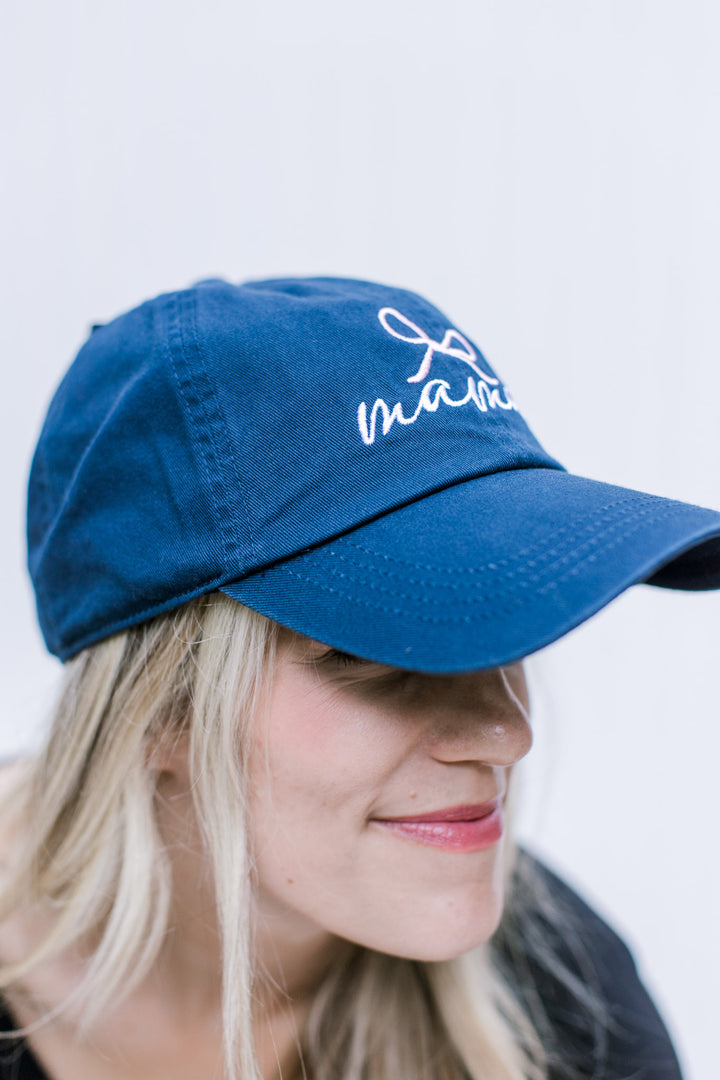 Side view of Model wearing an blue adjustable size hat with embroidered script mama with a pink bow.