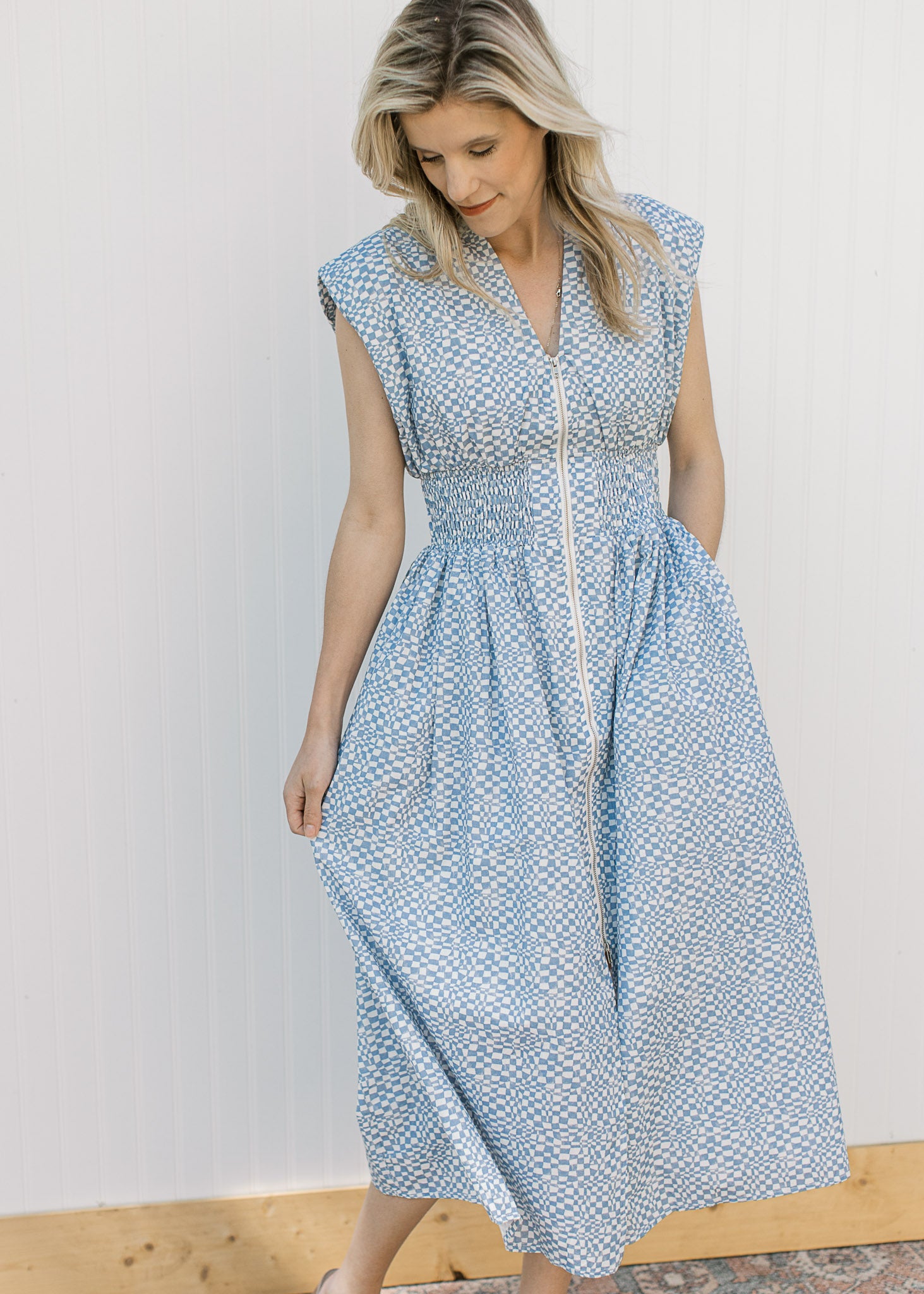 Model wearing a sleeveless blue and white checked midi with a front zipper and a smocked waist. 