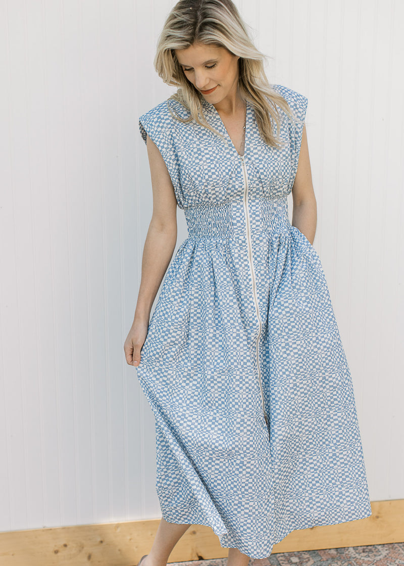 Model wearing a sleeveless blue and white checked midi with a front zipper and a smocked waist. 