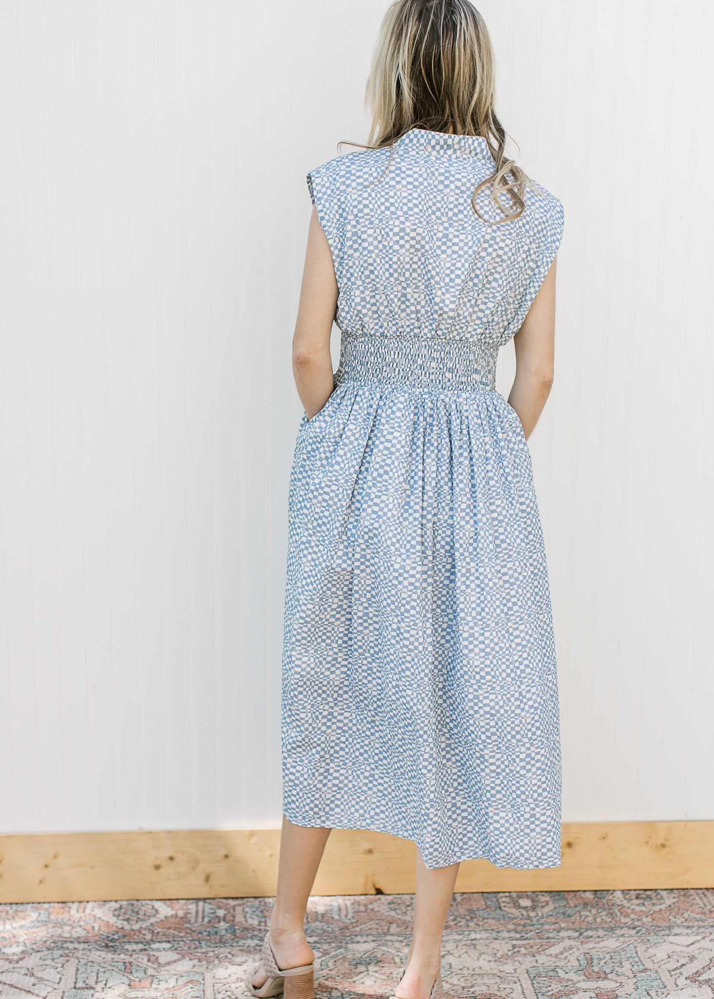 Back view of Model wearing a sleeveless blue and white checked midi with a front zipper. 