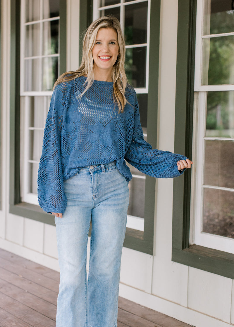 Model wearing a white shirt under a blue open weave sweater with stitched flowers and long sleeves.