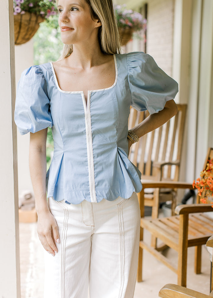 Model wearing a baby blue top with white piping at placket and scoop neck and bubble short sleeves.