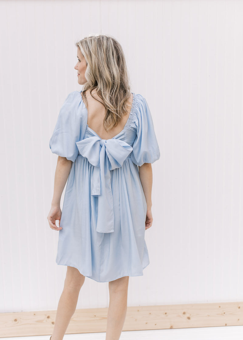 Back view of Model wearing a pale blue dress with a pleated bodice, square neck and short sleeves.