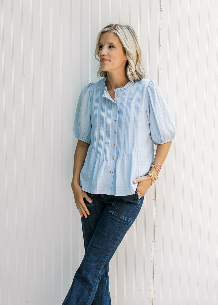 Model wearing jeans and a baby blue top with pleated front, round neck with ruffle and button-front.