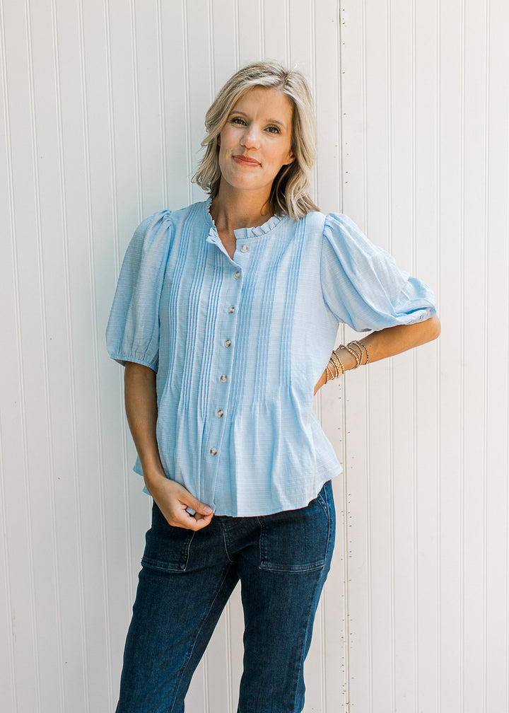 Model wearing a baby blue top with pleated front, round neck with ruffle and button-front.