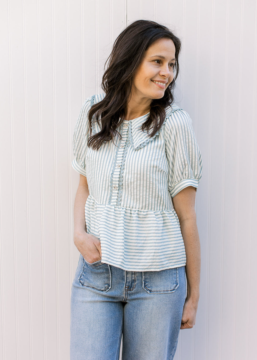 Model wearing a pale blue and cream striped top with a button placket with scalloped edges. 