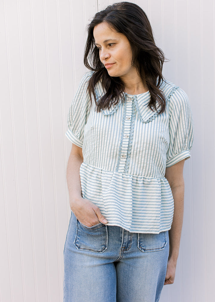 Model wearing a pale blue and cream striped peplum top with scalloped detail, short sleeves. 