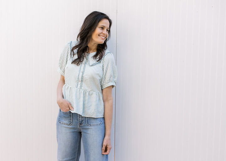 Model wearing a pale blue and cream striped peplum top with a button placket and short sleeves.