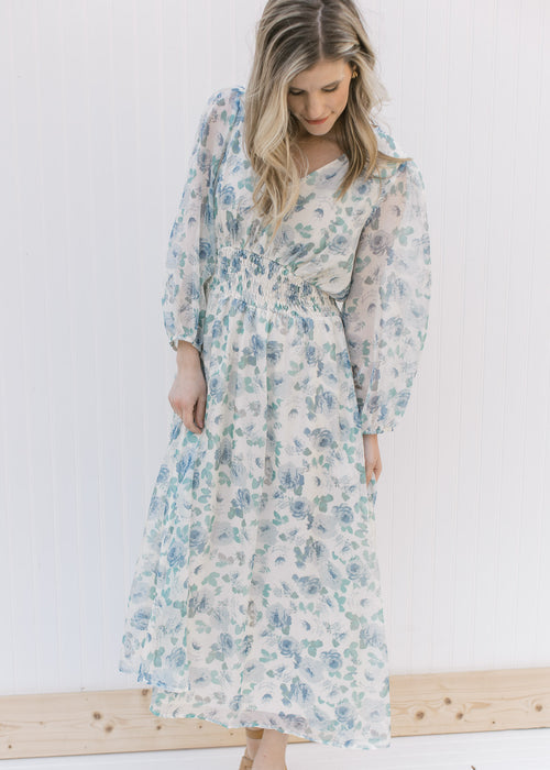 Model wearing a cream midi adorned with blue roses, an elastic waist and sheer 3/4 sleeves. 