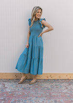 Model wearing flats and a dusty blue midi with a smocked bodice, flutter sleeves and tiered skirt.