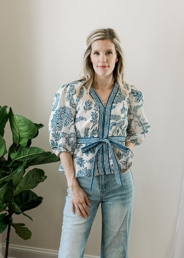 Model wearing jeans and a cream v-neck top with blue print, 3/4 sleeves and blue stripe detail. 