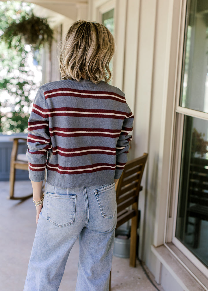 Back view of Model wearing a blue cardigan with maroon and cream stripes, buttons and long sleeves. 