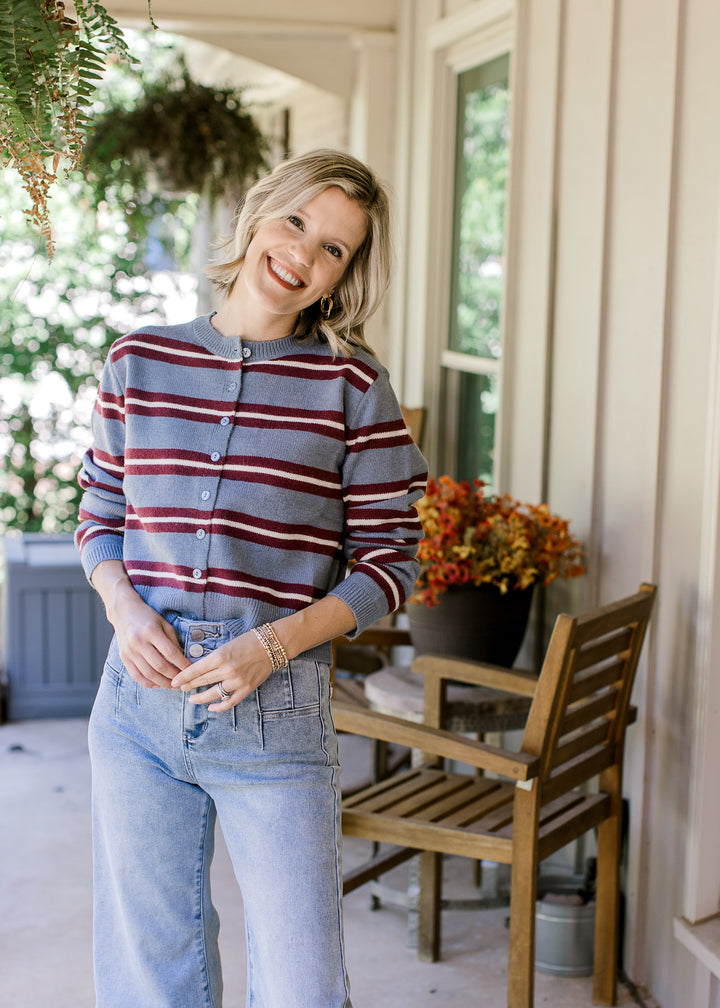 Model wearing jeans and a blue cardigan with maroon and cream stripes, buttons and long sleeves. 