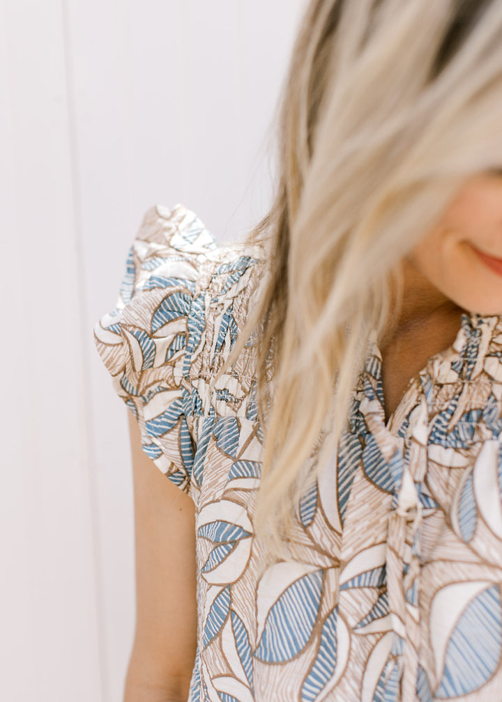 Close up of ruffle cap sleeve and v-neck with a smocked yolk on a cream, tan and blue top. 