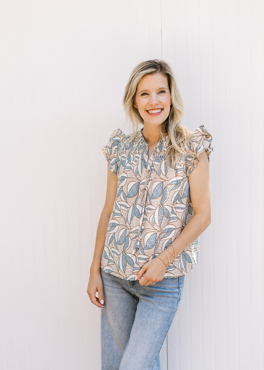 Model wearing a lightweight top with tan, cream and blue pattern, v-neck and ruffle cap sleeves. 