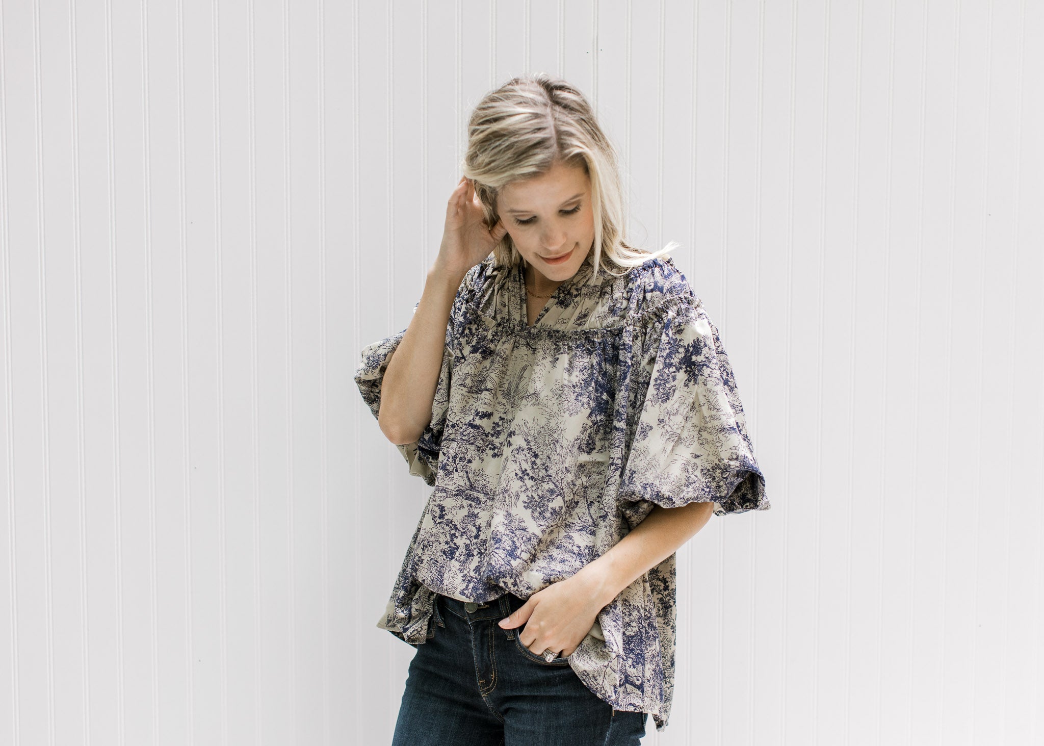 Model wearing a tan v-neck top with navy toile print, bubble short sleeves and ruched neck detail. 