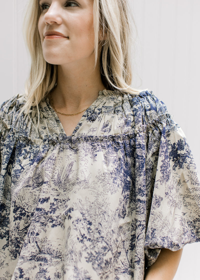 Close up of navy toile print on a tan top with a ruched v-neck and bubble short sleeves. 